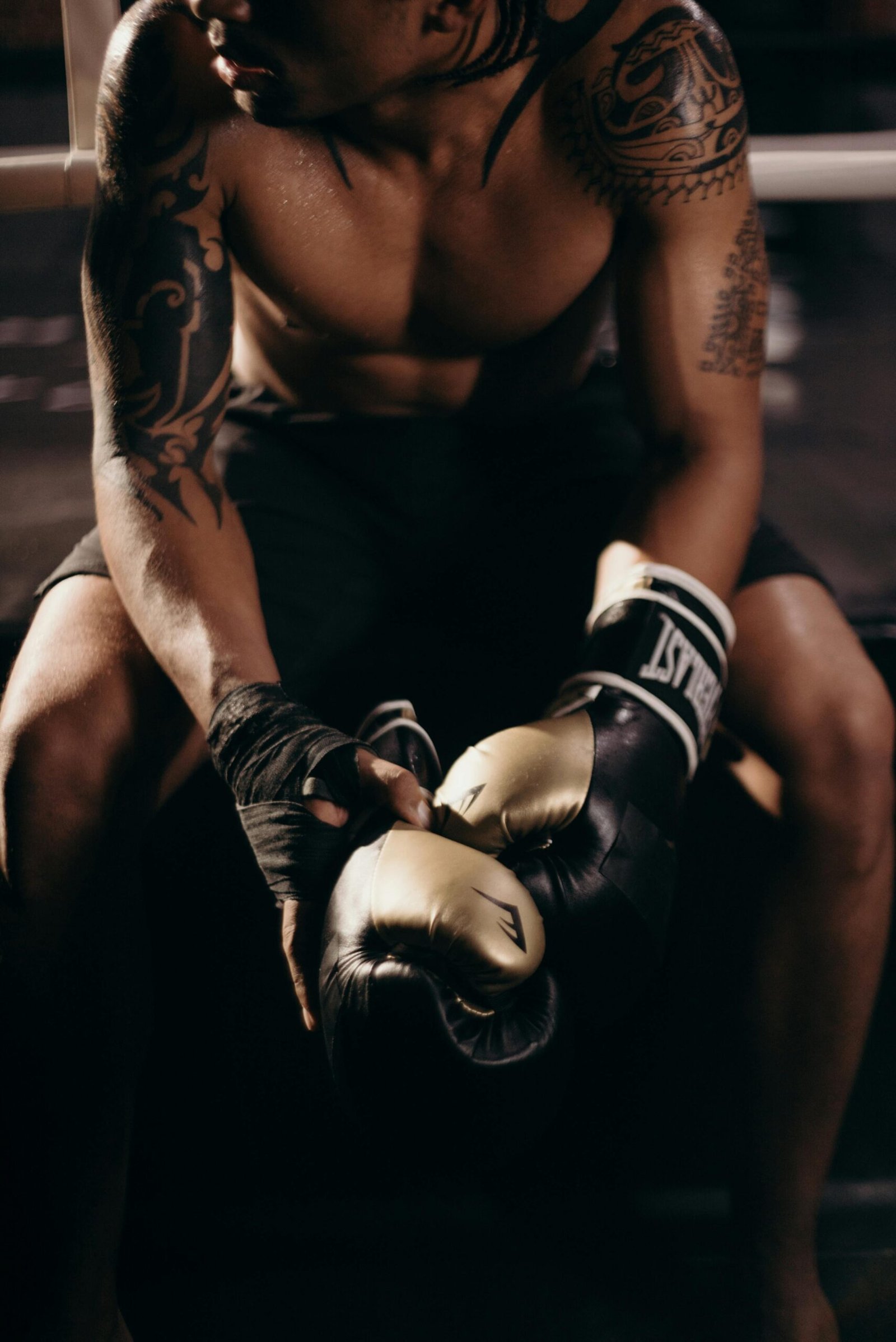 A muscular boxer with tattoos sitting shirtless in a gym holding boxing gloves.